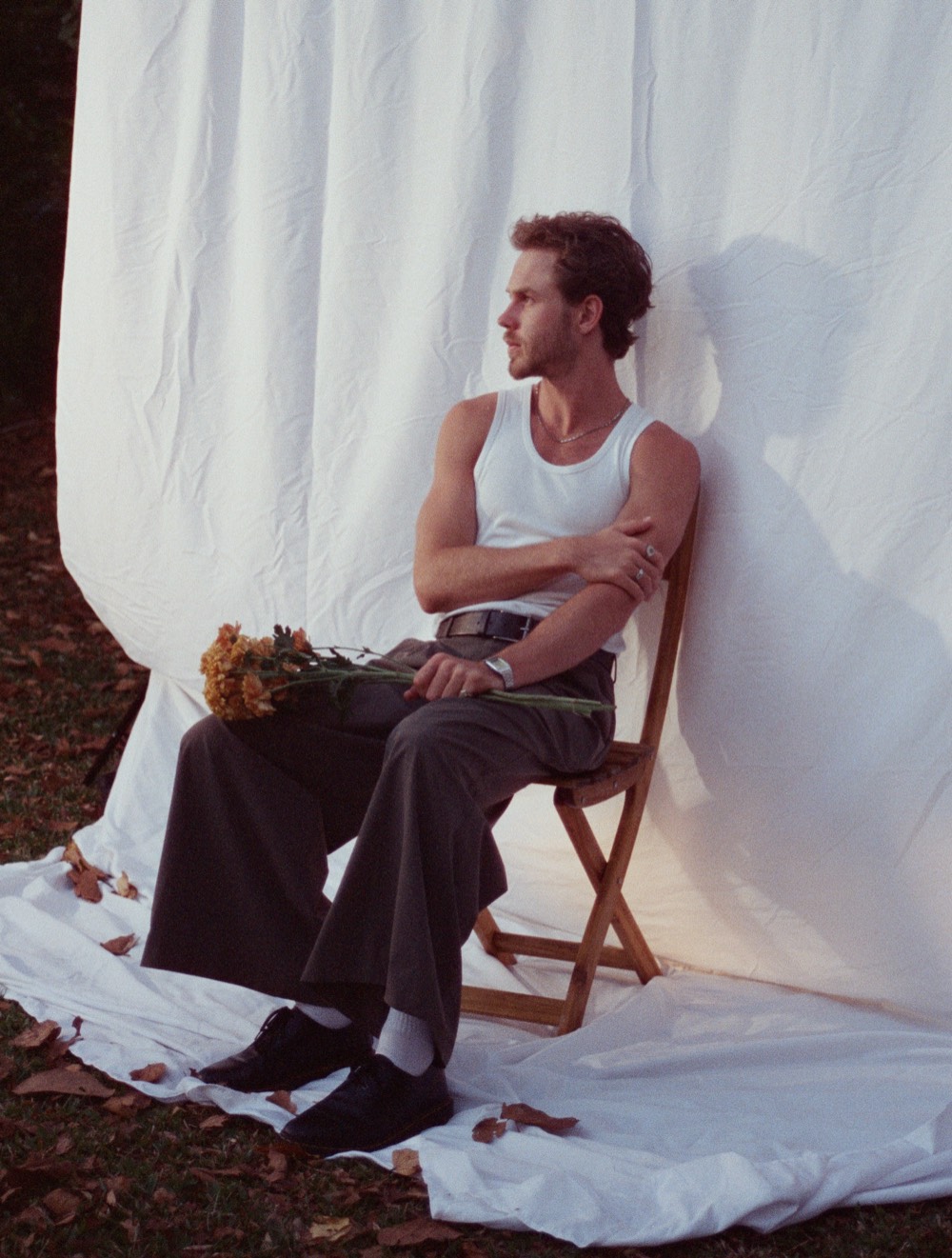 Henry West seated on a wooden chair against a white sheet hung outdoors, holding orange flowers, looking away.