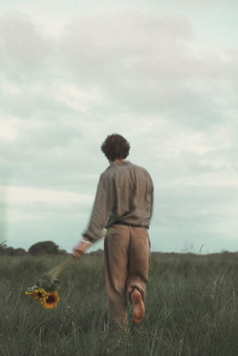 Henry West walking barefoot away from the camera through long grass, sunflowers trailing from one hand.