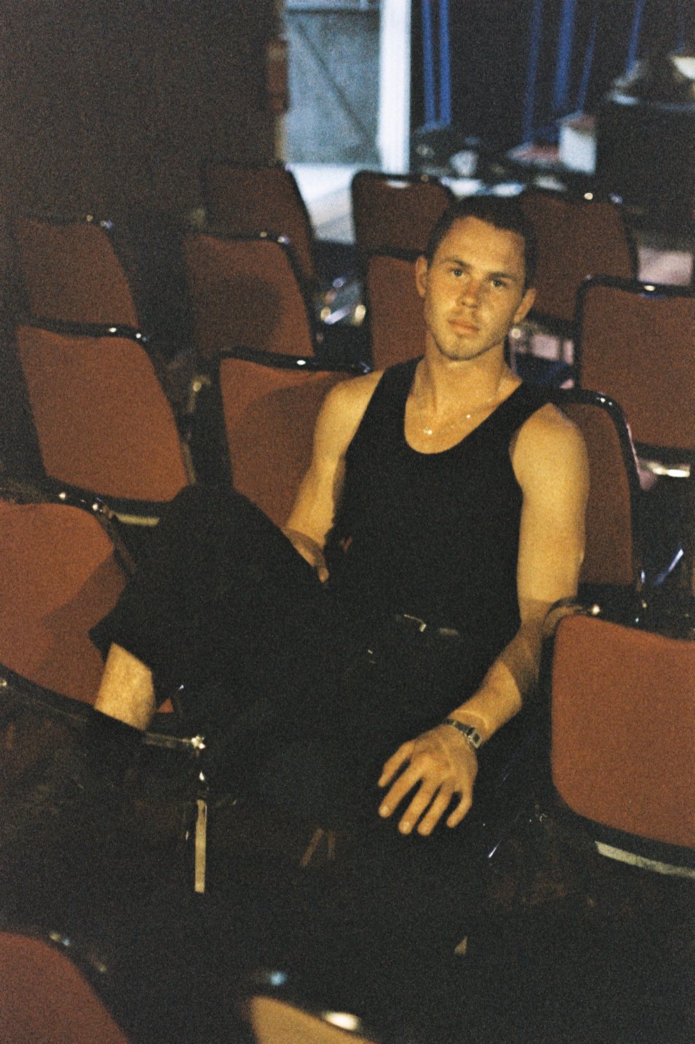 Henry West in a black singlet, leaning back across rows of red theatre seats, looking at the camera.